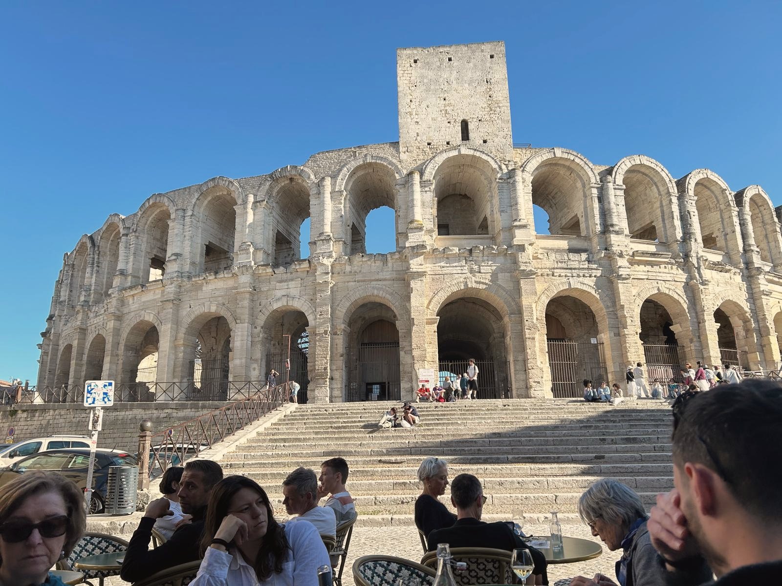 The Roman arena of Arles under a clear blue sky, with people at café tables in front