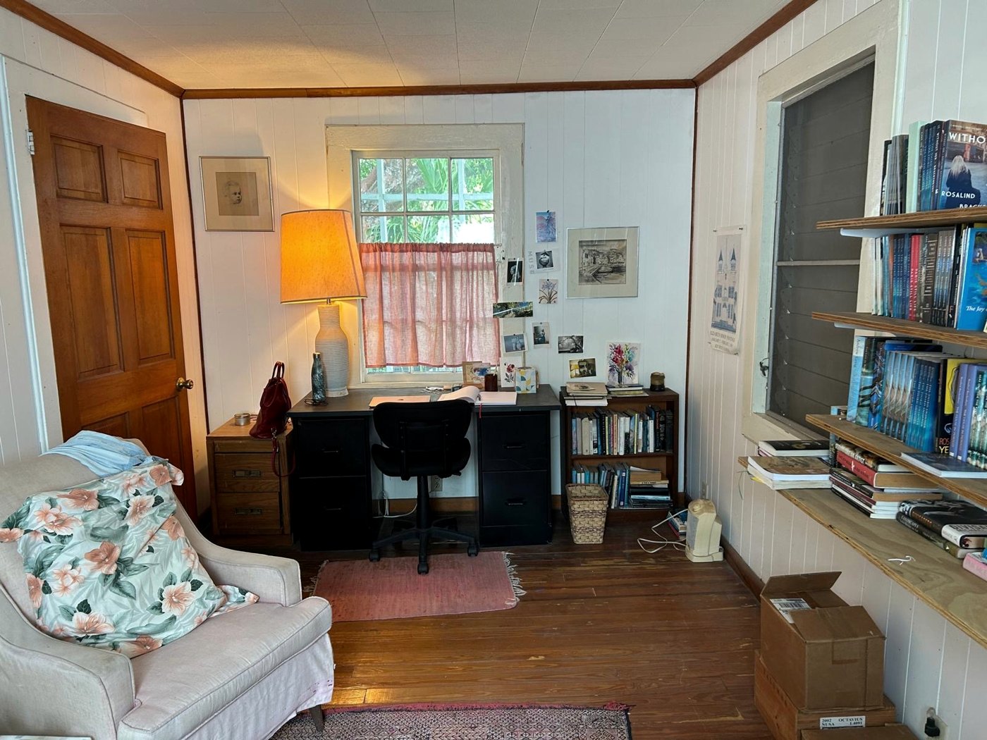 Ros's writing desk beneath a curtained window in her Key West studio, a lamp lit beside it, shelves of books along the wall