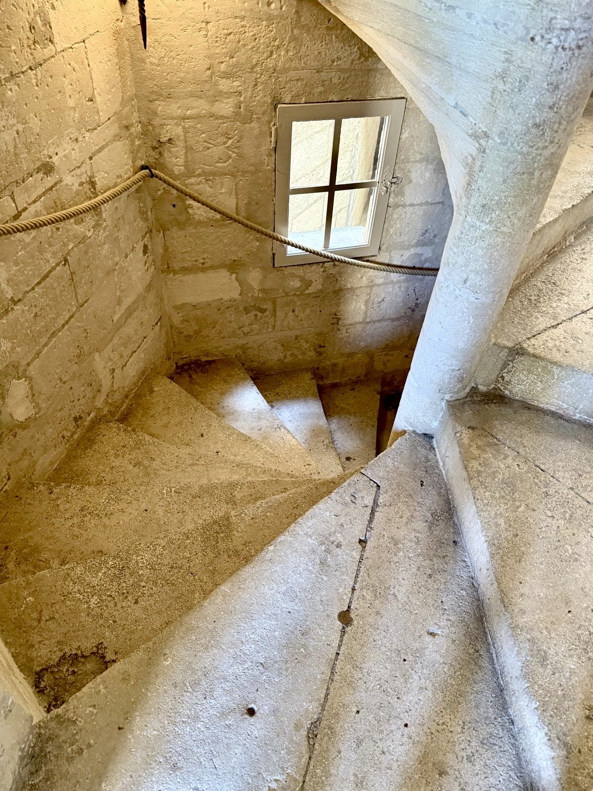 A worn stone spiral staircase with a rope handrail and a small window