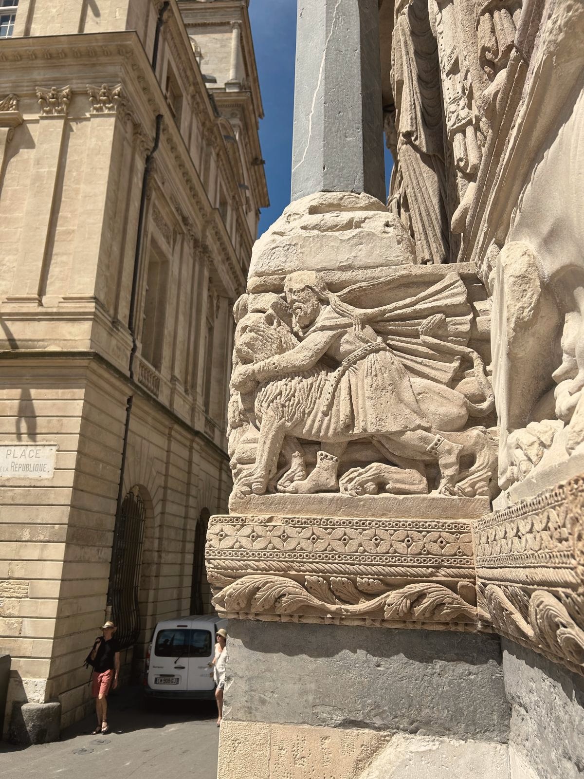 Carved Romanesque stone figures on a column against pale Arles buildings