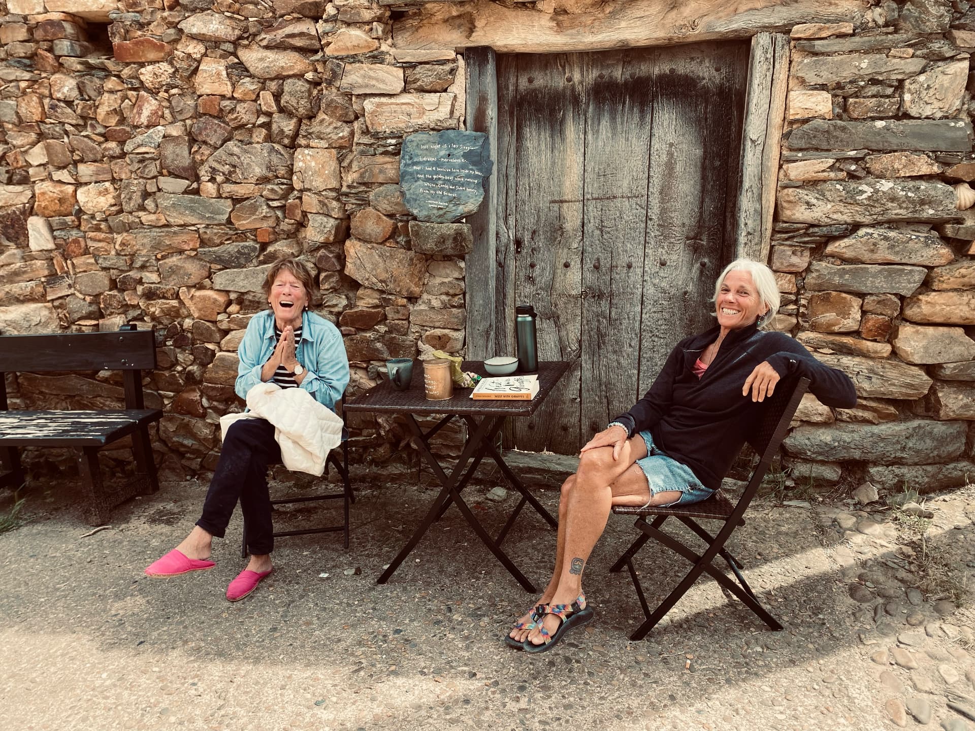 Rosalind Brackenbury and her friend Carol laughing together before a stone wall bearing a painted plaque of Antonio Machado’s lines
