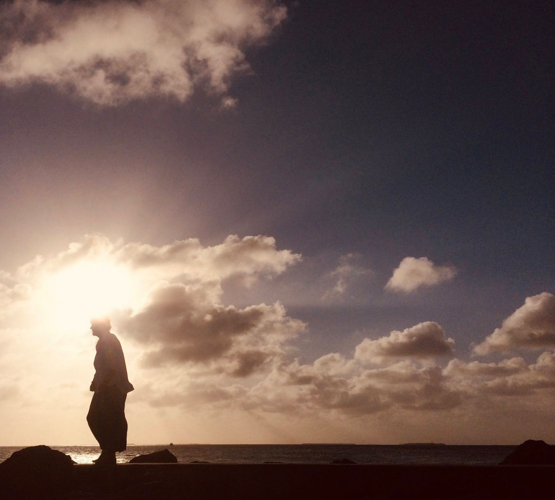 A figure in silhouette at the edge of the sea beneath evening clouds