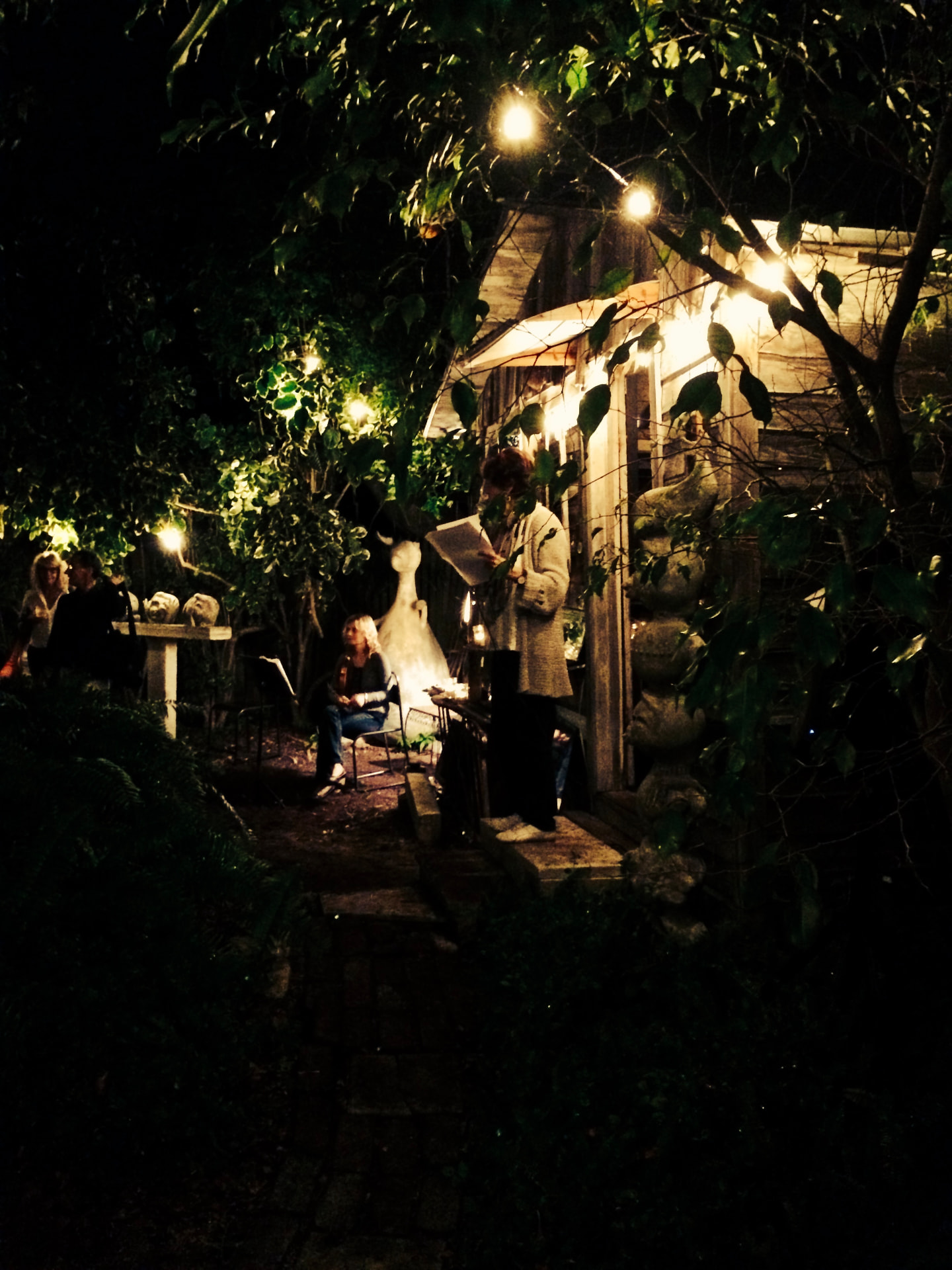 Ros reading aloud from a sheet of paper in a garden strung with lights