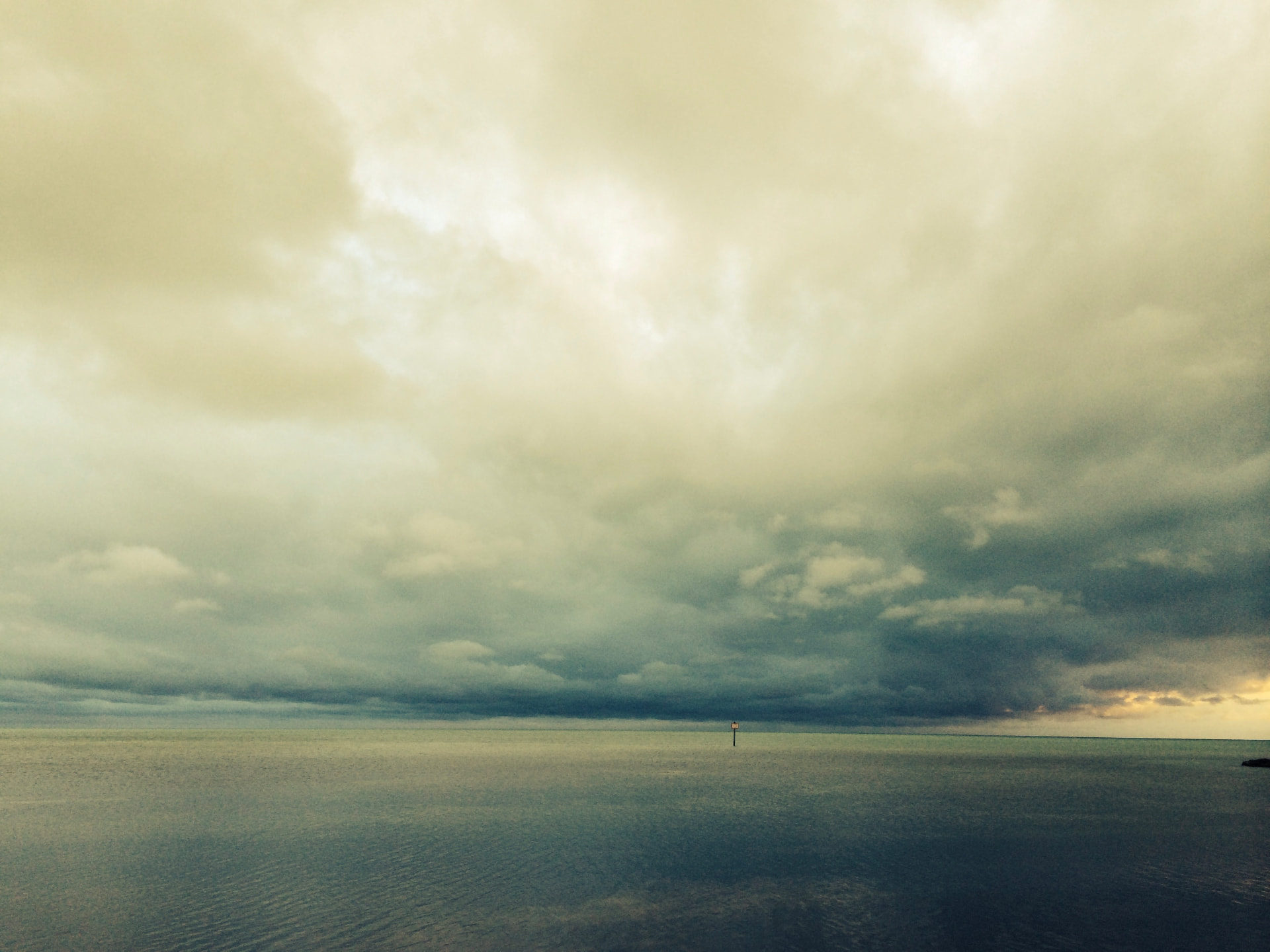 A wide sea horizon beneath dark, layered storm clouds, with a lone marker in the distance