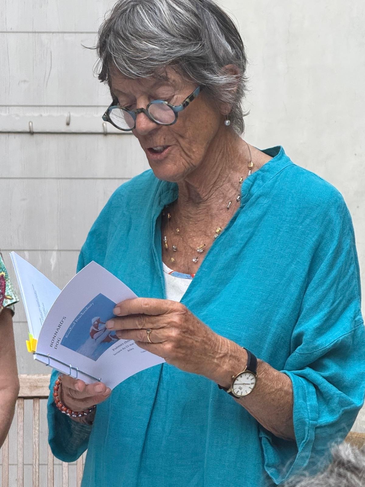 Rosalind Brackenbury in a turquoise linen shirt reading from a booklet of her poems