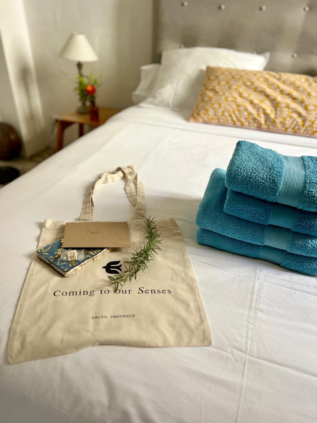 A Coming to Our Senses tote bag with a sprig of rosemary on a bed beside turquoise towels