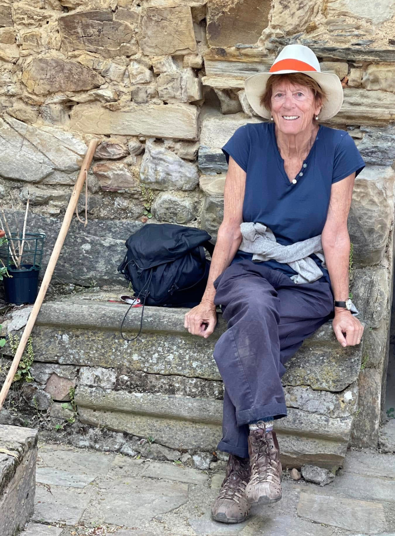 Rosalind Brackenbury resting on a stone bench in hiking boots and a sun hat, a walking stick beside her
